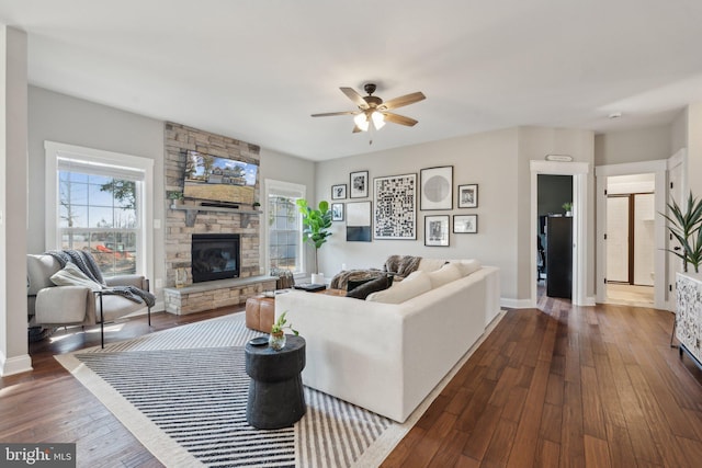 living area featuring ceiling fan, plenty of natural light, a stone fireplace, and dark wood-style floors