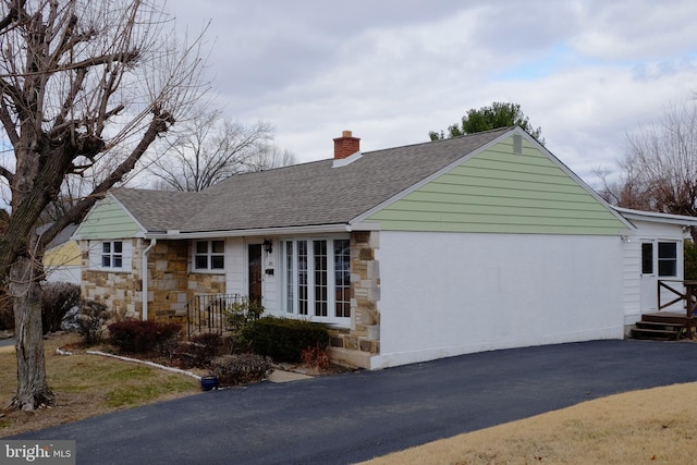 ranch-style house with stone siding, a shingled roof, a chimney, and aphalt driveway