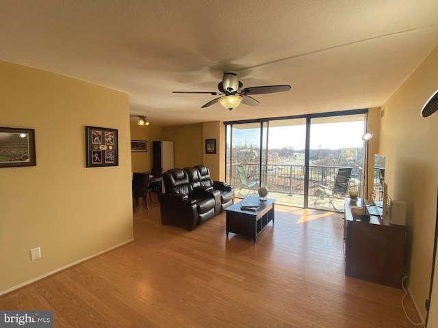 living area with a ceiling fan, a textured ceiling, wood finished floors, a wall of windows, and baseboards