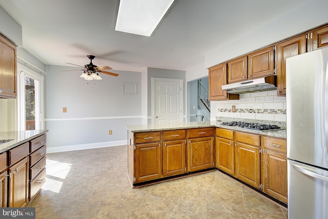 kitchen with brown cabinetry, a peninsula, decorative backsplash, under cabinet range hood, and appliances with stainless steel finishes