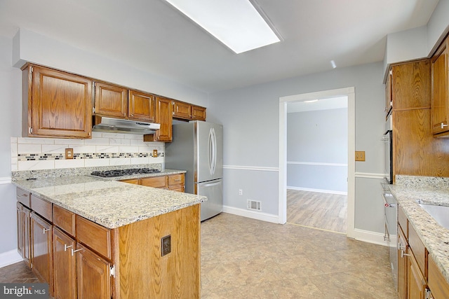 kitchen featuring brown cabinets, gas stovetop, freestanding refrigerator, under cabinet range hood, and tasteful backsplash