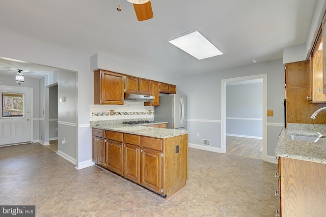 kitchen featuring a peninsula, freestanding refrigerator, a sink, under cabinet range hood, and backsplash