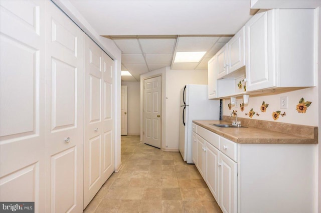 kitchen with a sink, a drop ceiling, freestanding refrigerator, and white cabinetry