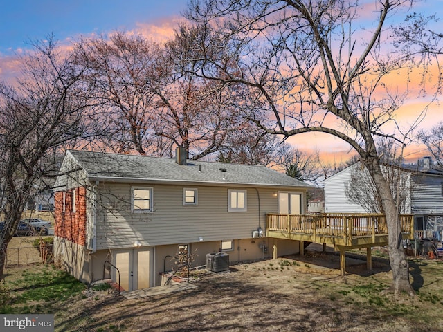 back of house at dusk featuring a deck, central air condition unit, a shingled roof, and a chimney