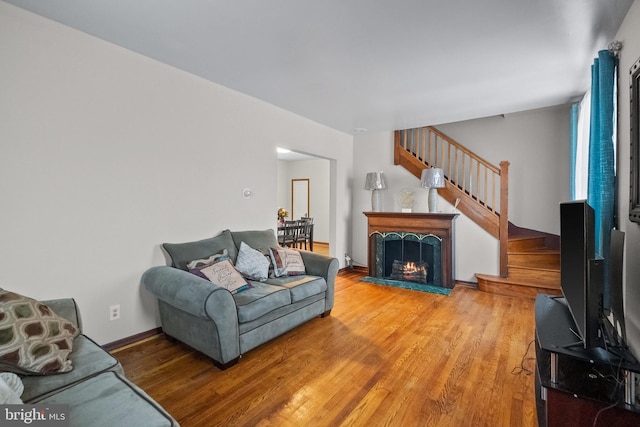 living room featuring light wood finished floors, a warm lit fireplace, stairs, and baseboards