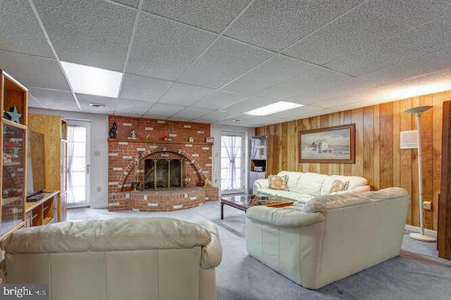 living room featuring a brick fireplace, carpet, a drop ceiling, and wood walls