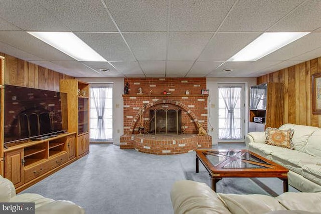 carpeted living room with a brick fireplace, a drop ceiling, and wood walls