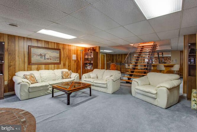 carpeted living area with a paneled ceiling, visible vents, wooden walls, and stairway