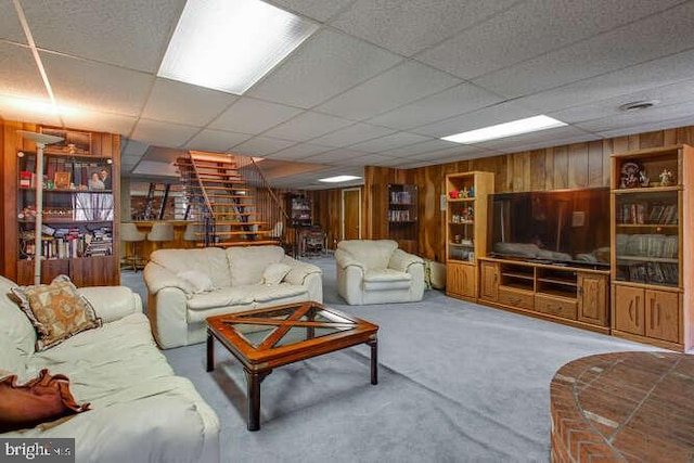 carpeted living room featuring a drop ceiling, wooden walls, and stairs