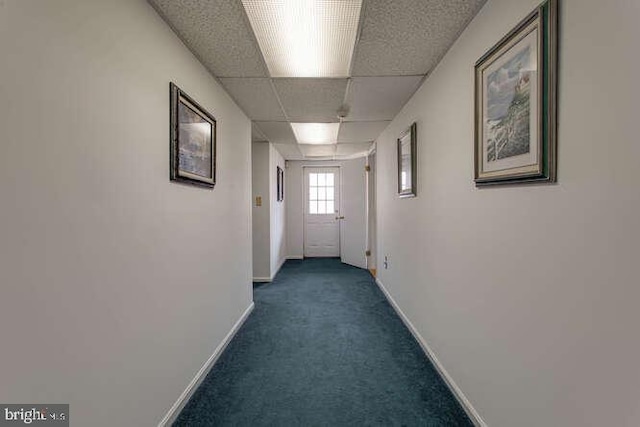 hall with dark colored carpet, a drop ceiling, and baseboards