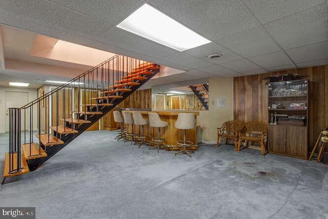 bar featuring a paneled ceiling, wooden walls, concrete floors, stairway, and a bar