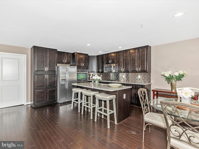 kitchen with dark wood-style floors, stainless steel appliances, dark brown cabinets, a kitchen breakfast bar, and tasteful backsplash