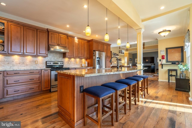 kitchen with ornamental molding, under cabinet range hood, decorative columns, stainless steel appliances, and decorative backsplash