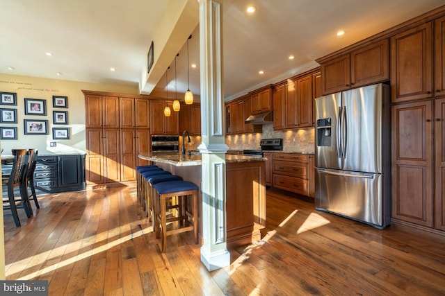 kitchen featuring brown cabinetry, a center island with sink, dark stone counters, dark wood-type flooring, and appliances with stainless steel finishes