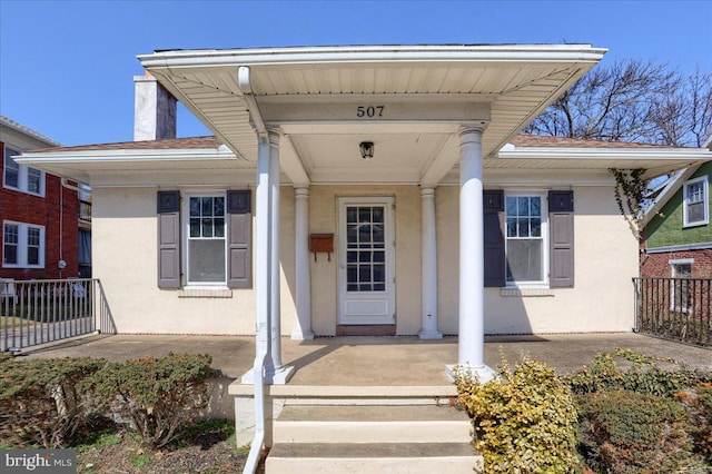 view of exterior entry with stucco siding, covered porch, a chimney, and fence