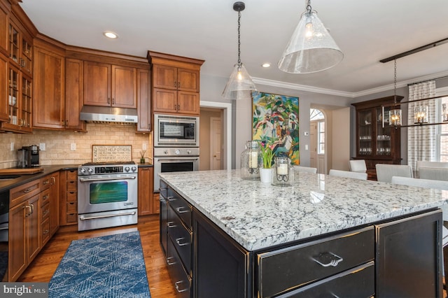 kitchen with under cabinet range hood, wood finished floors, appliances with stainless steel finishes, backsplash, and brown cabinetry