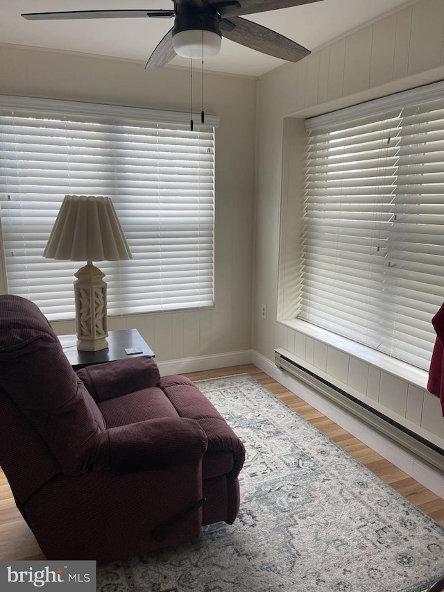 living area with a baseboard heating unit, ceiling fan, wood finished floors, and baseboards