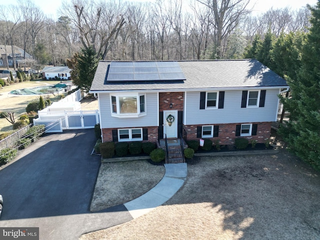 raised ranch featuring a gate, fence, roof with shingles, brick siding, and roof mounted solar panels