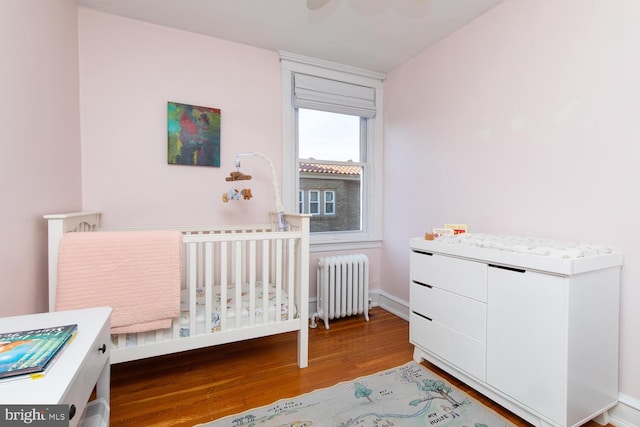 bedroom with radiator, wood finished floors, baseboards, ceiling fan, and a nursery area