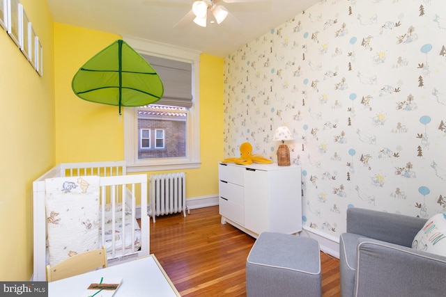 bedroom featuring radiator, wallpapered walls, baseboards, wood finished floors, and a nursery area