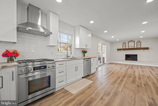 kitchen featuring a fireplace, a sink, light wood-style floors, appliances with stainless steel finishes, and wall chimney exhaust hood