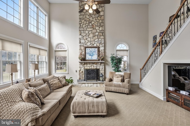 carpeted living room with baseboards, ceiling fan, a high ceiling, stairs, and a stone fireplace