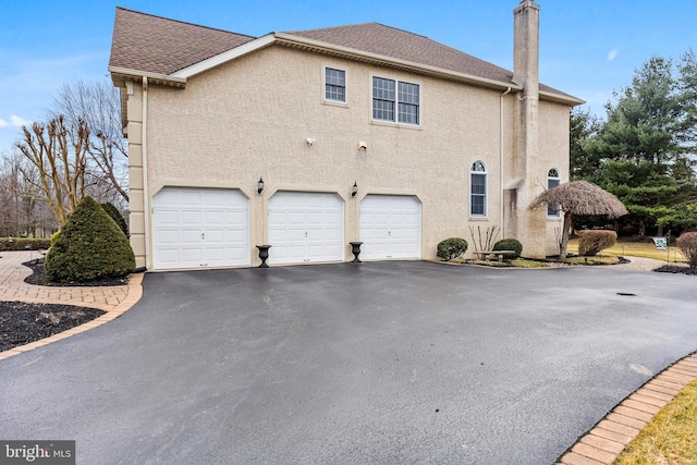 view of side of property featuring driveway, a shingled roof, a chimney, an attached garage, and stucco siding