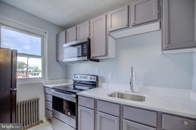 kitchen with radiator heating unit, light stone counters, stainless steel appliances, gray cabinetry, and a sink