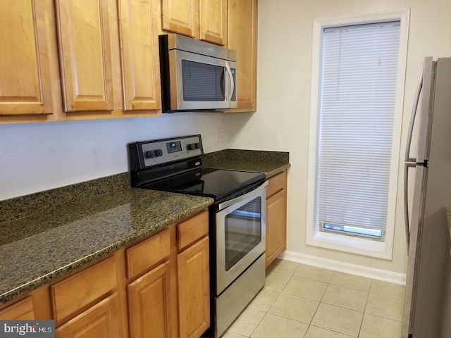 kitchen with light tile patterned flooring, baseboards, appliances with stainless steel finishes, brown cabinets, and dark stone counters