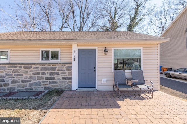 doorway to property featuring a patio area, stone siding, and a shingled roof