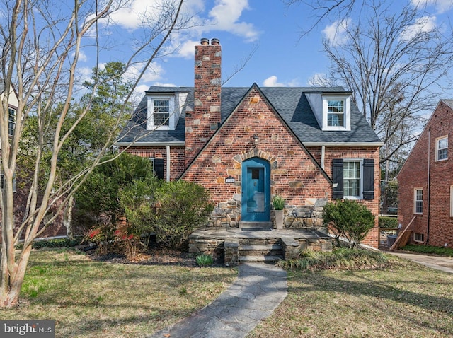 tudor-style house featuring a front lawn, brick siding, roof with shingles, and a chimney