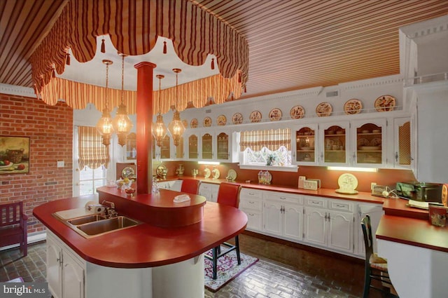 kitchen featuring brick floor, a kitchen island with sink, brick wall, a sink, and white cabinets