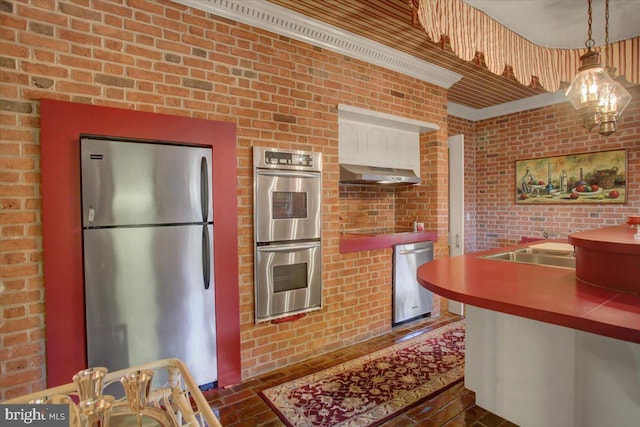 kitchen featuring brick floor, under cabinet range hood, stainless steel appliances, brick wall, and hanging light fixtures