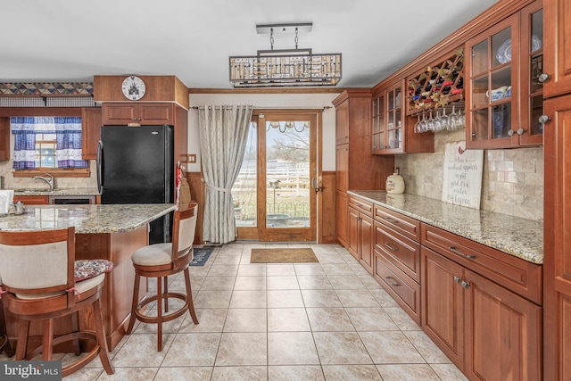 kitchen featuring freestanding refrigerator, a breakfast bar area, light tile patterned flooring, brown cabinetry, and glass insert cabinets