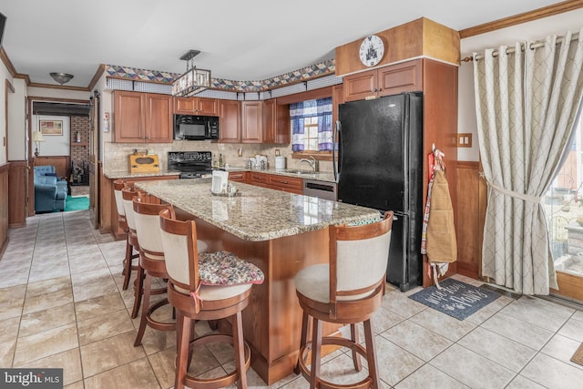 kitchen featuring crown molding, light stone counters, wainscoting, brown cabinetry, and black appliances