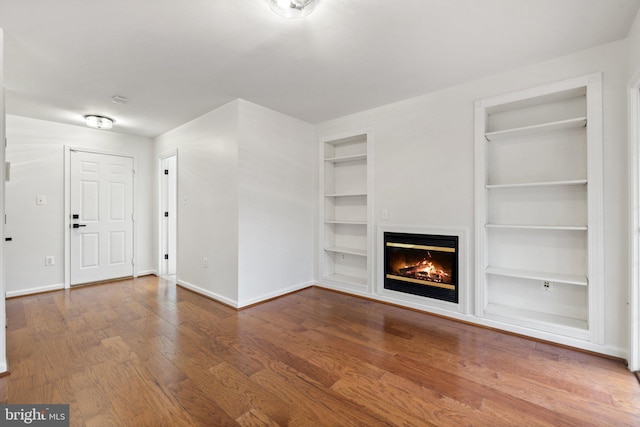 unfurnished living room featuring a glass covered fireplace, wood finished floors, built in shelves, and baseboards