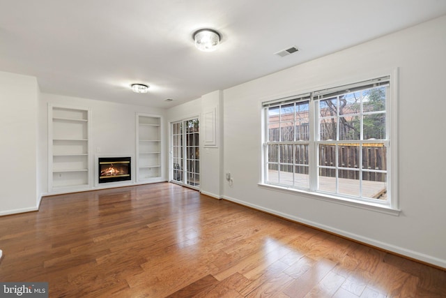 unfurnished living room featuring built in shelves, visible vents, baseboards, hardwood / wood-style floors, and a glass covered fireplace