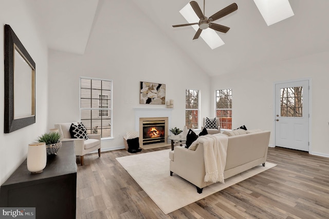 living area featuring wood finished floors, high vaulted ceiling, a skylight, a fireplace with flush hearth, and ceiling fan