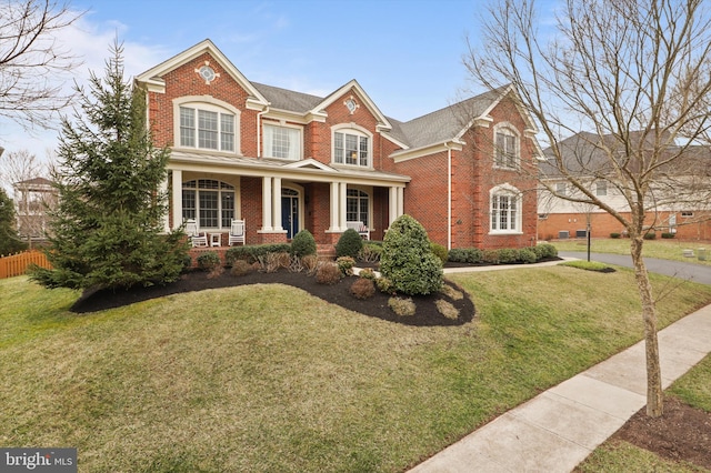 traditional home with a front yard, fence, roof with shingles, a porch, and brick siding