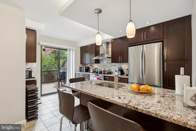 kitchen featuring dark brown cabinetry, stainless steel appliances, a sink, wall chimney exhaust hood, and a kitchen bar