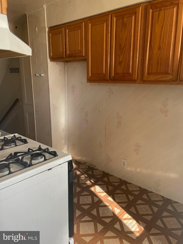 kitchen with white gas stove, range hood, brown cabinetry, light countertops, and light floors