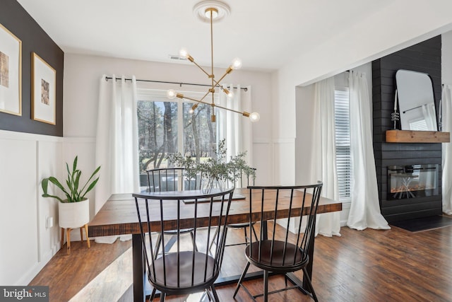 dining area featuring dark wood-style floors, a fireplace, a notable chandelier, a decorative wall, and wainscoting