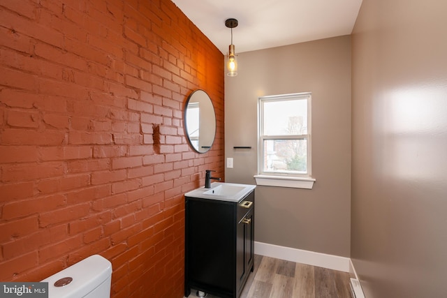 half bath featuring toilet, brick wall, wood finished floors, vanity, and baseboards