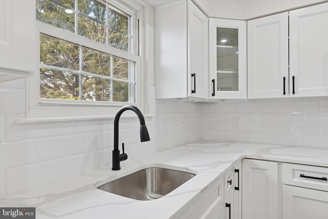 kitchen with light stone counters, tasteful backsplash, glass insert cabinets, white cabinetry, and a sink