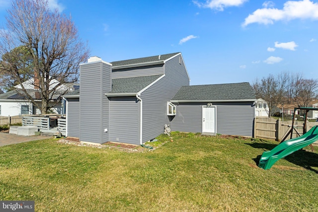 rear view of house with fence, a yard, a chimney, a shingled roof, and a playground