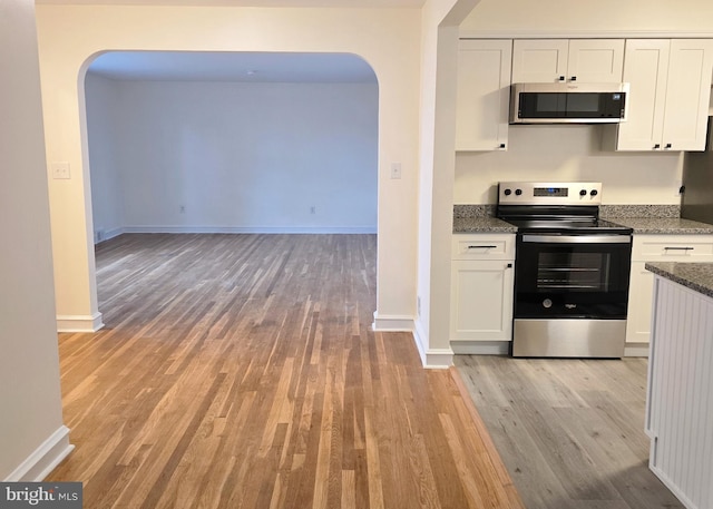 kitchen with dark stone counters, arched walkways, stainless steel appliances, white cabinets, and light wood-style floors