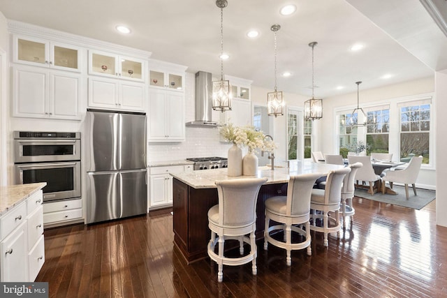 kitchen with stainless steel appliances, light stone counters, wall chimney exhaust hood, and a center island with sink