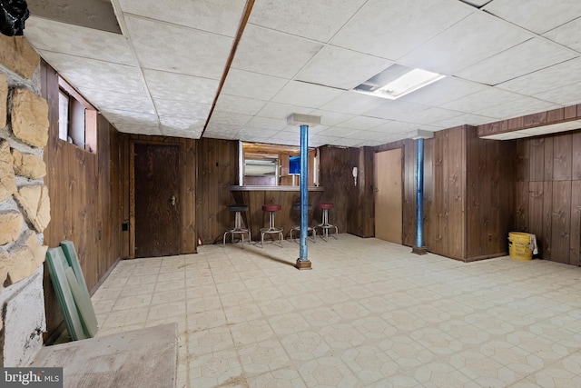 finished below grade area featuring tile patterned floors, a drop ceiling, wooden walls, and a bar