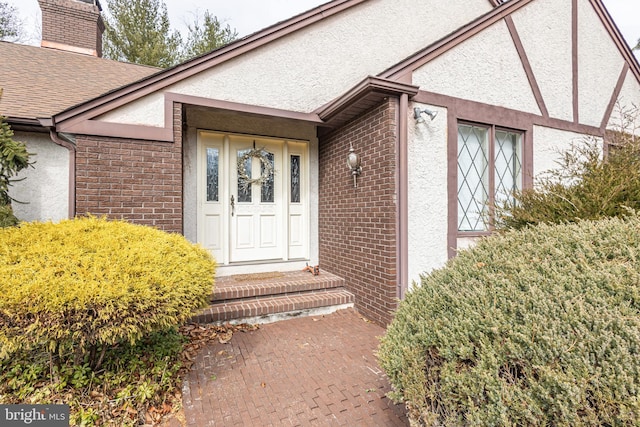 view of exterior entry with a shingled roof, a chimney, brick siding, and stucco siding