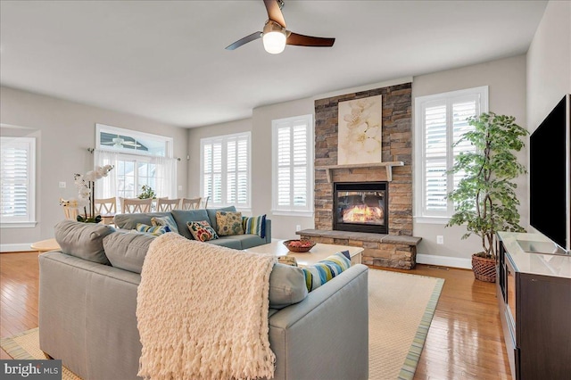 living area with ceiling fan, a stone fireplace, wood-type flooring, and baseboards